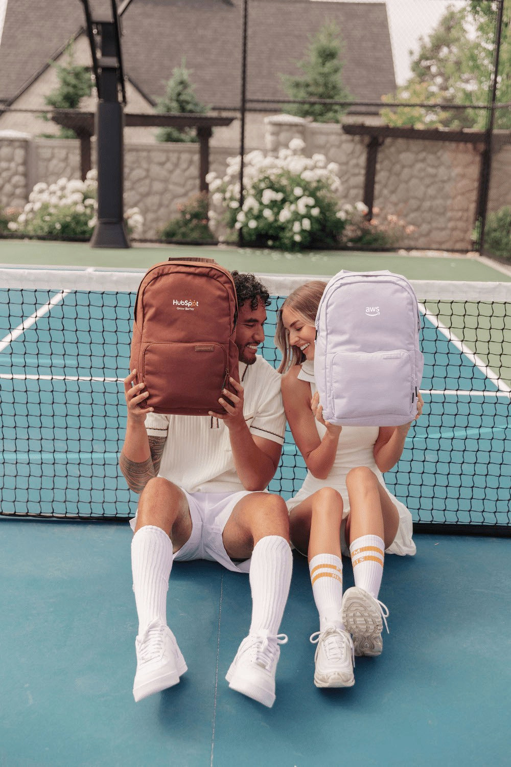 Two people sitting on a tennis court holding Brevitē backpacks embroidered with the HubSpot and AWS logos
