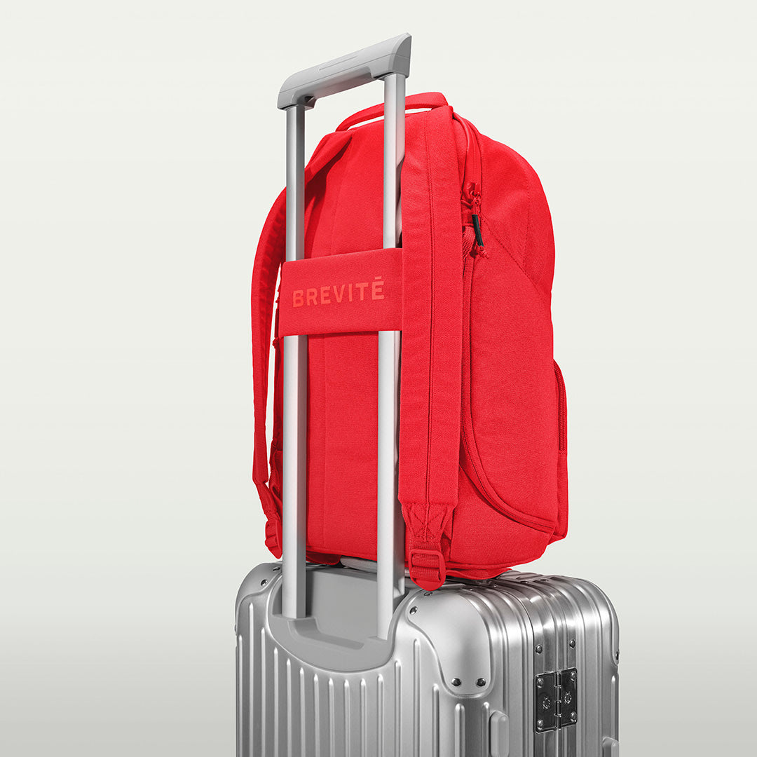 The Brevitē Backpack in Poppy Red, luggage strap over a suitcase handle
