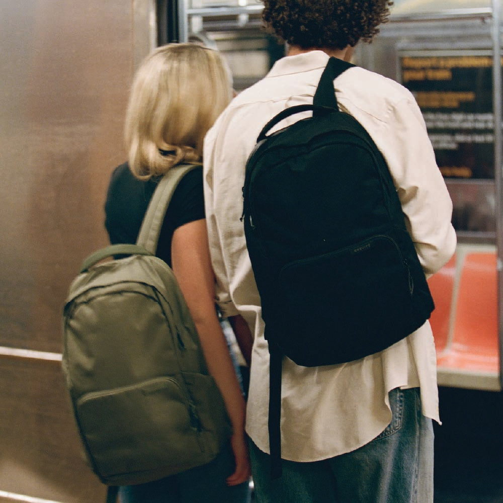 Two commuters on a subway platform wearing olive and black Brevitē backpacks