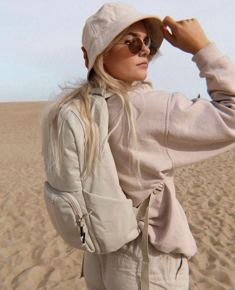 A person in casual attire and sunglasses stands in a desert wearing a backpack and a bucket hat.