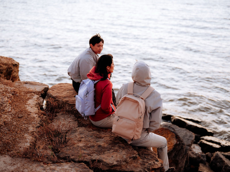 Three friends sitting on rocks by the water, two wearing Brevitē backpacks