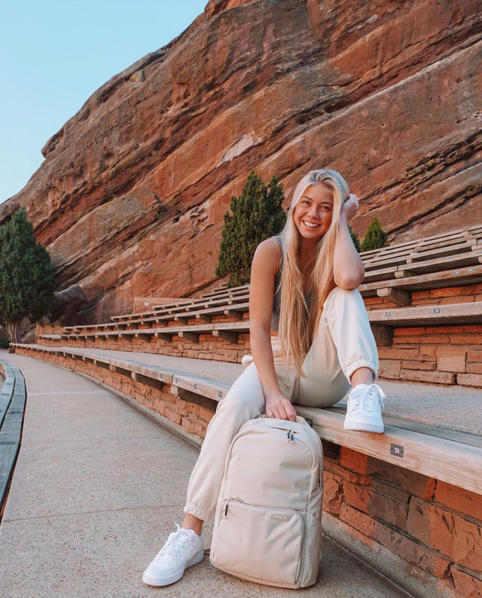 A woman sitting on the benches at Red Rocks Amphitheatre with a cream Brevitē backpack