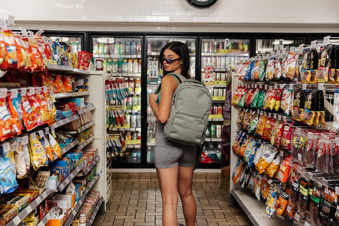 A woman wearing a sage Brevitē backpack in a convenience store aisle