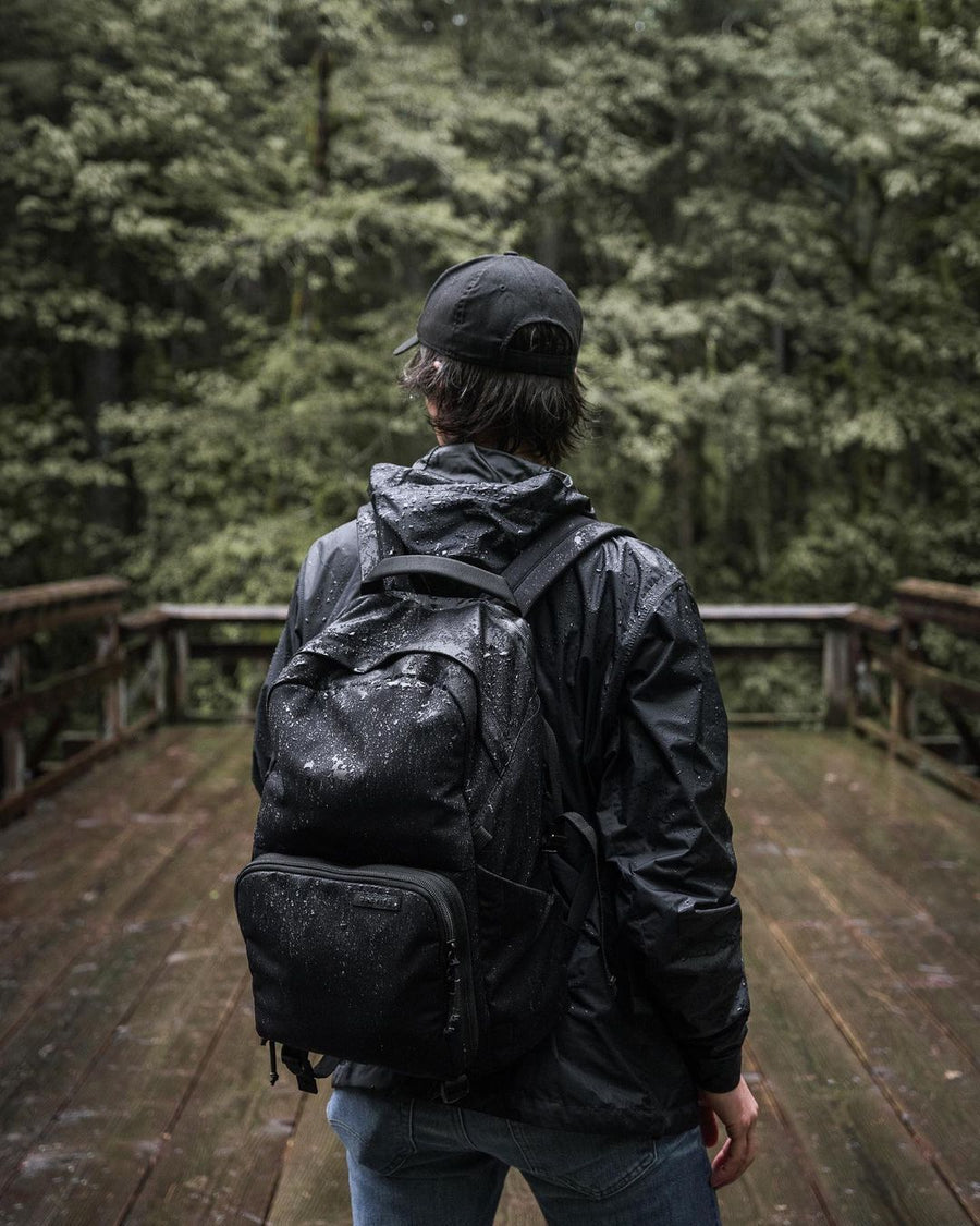A man in a rain jacket on a wooden deck in a forest, rain beading on his black Brevitē backpack