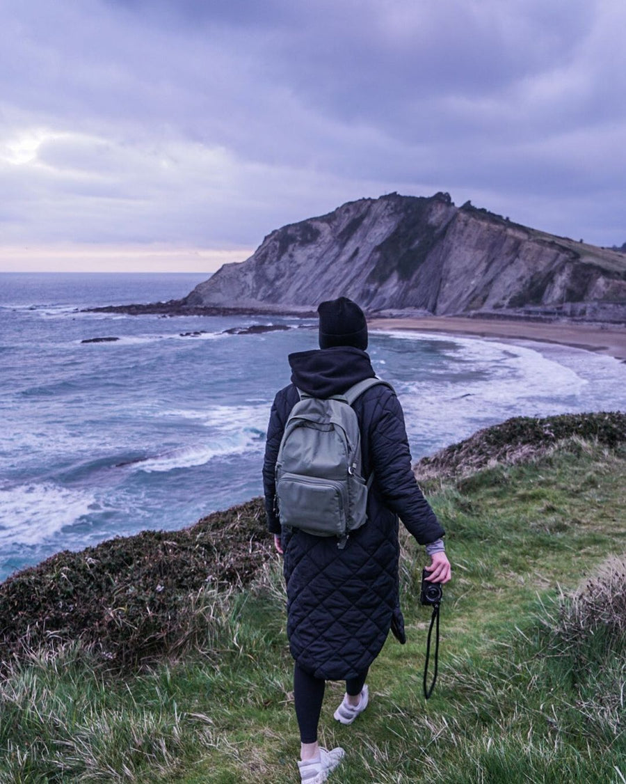 A person in a long black coat walking a cliff path above the ocean, camera in hand and a green Brevitē backpack on