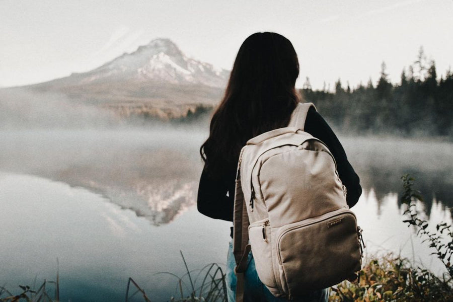 A woman with a cream Brevitē backpack at a misty lake facing a snow-capped mountain
