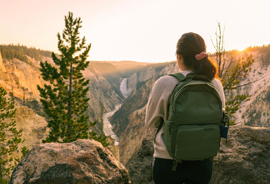 A woman with a green Brevitē backpack watching sunrise over a canyon waterfall