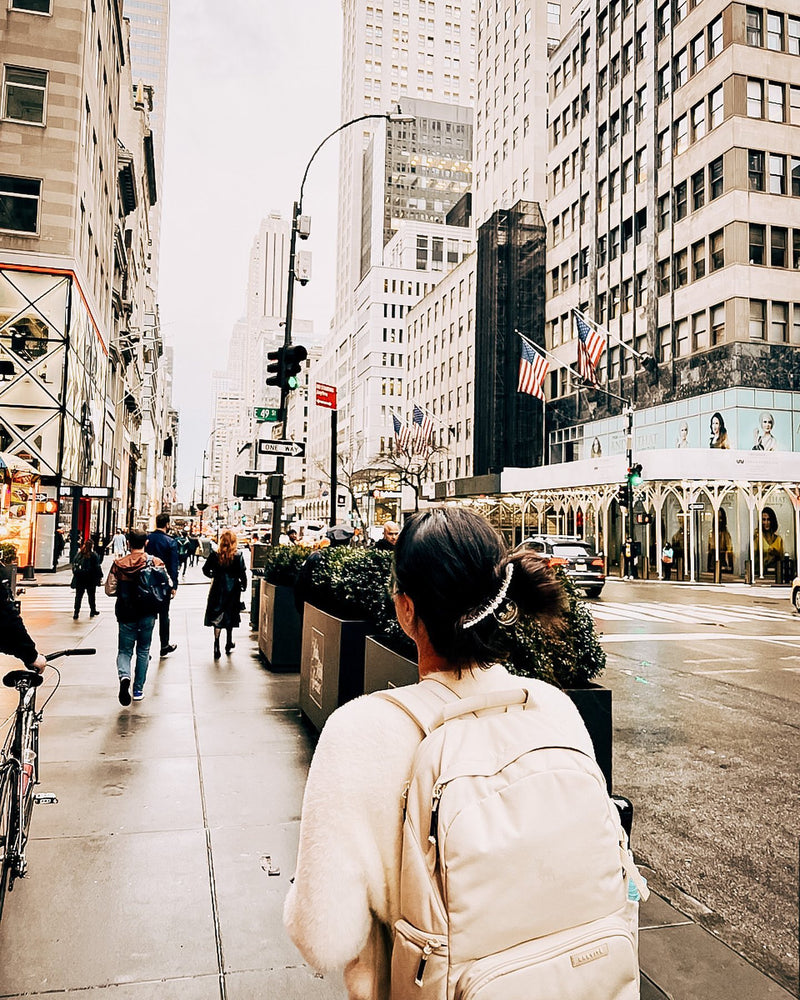 A woman with a cream Brevitē backpack walking down a busy Manhattan sidewalk