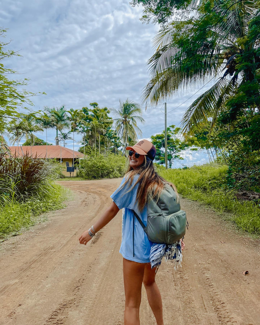 A smiling woman in a trucker hat with a green Brevitē backpack on a dirt road lined with palm trees