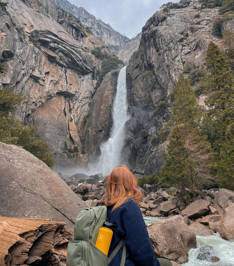 A woman with a green Brevitē backpack and a water bottle in the side pocket looking up at a waterfall