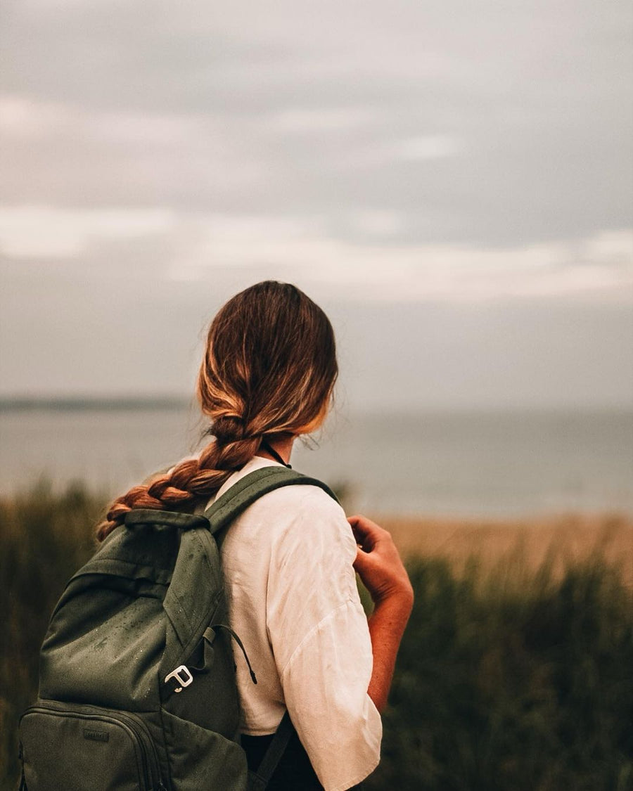 A woman with a long braid and a green Brevitē backpack looking out at a gray sea