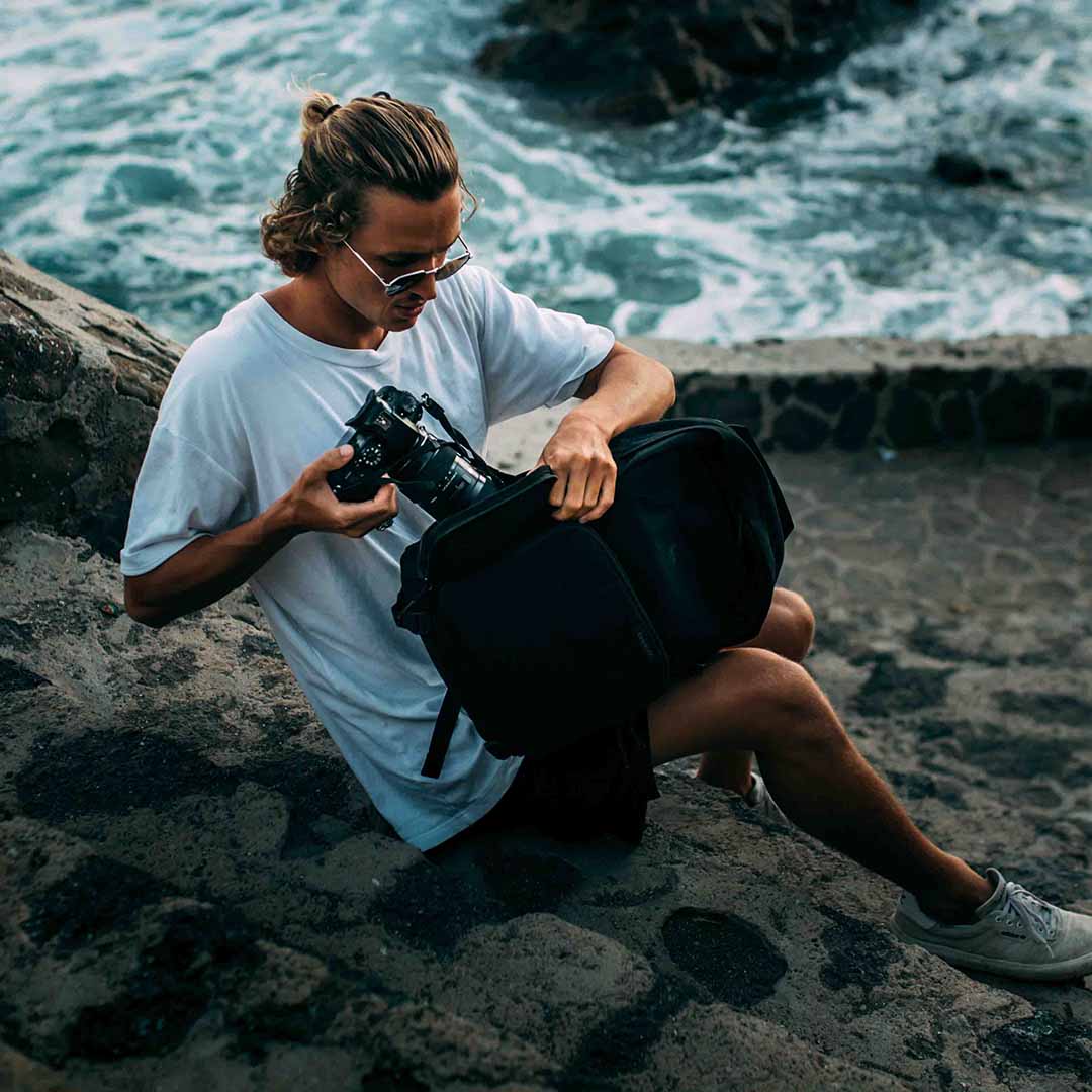 A photographer on a rocky shoreline reaching into a black Brevitē Jumper for his camera